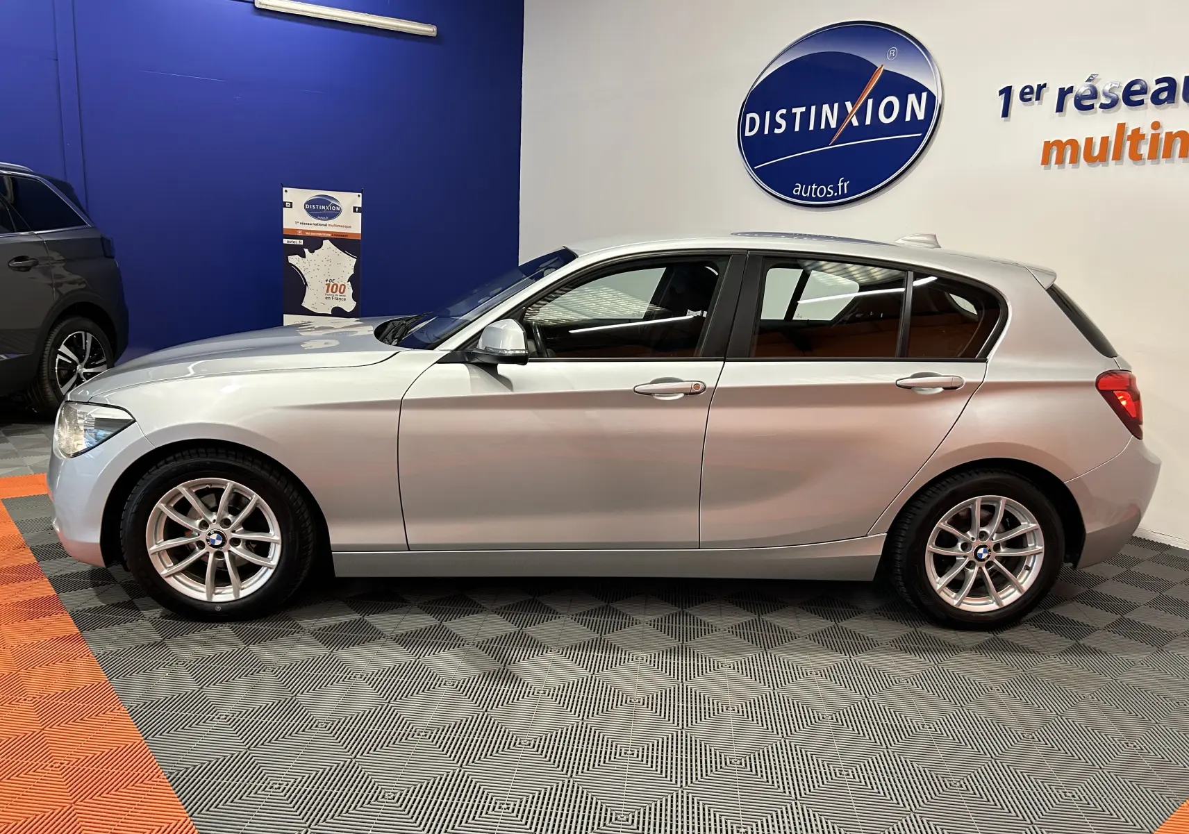 Profil côté gauche d'une BMW Série 1 gris clair 5 portes, stationnée en intérieur sur sol damier, avec jantes alliage visibles.