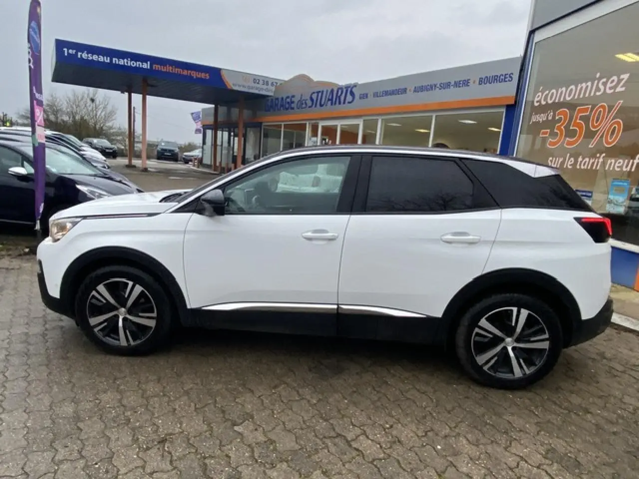 Profil côté gauche du Peugeot 3008 blanc avec toit noir et jantes alliage 18 pouces, stationné devant un garage.