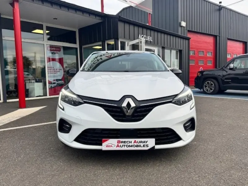 Renault Clio blanc glacier vue de face, avec calandre noire et phares LED, devant un garage automobile.