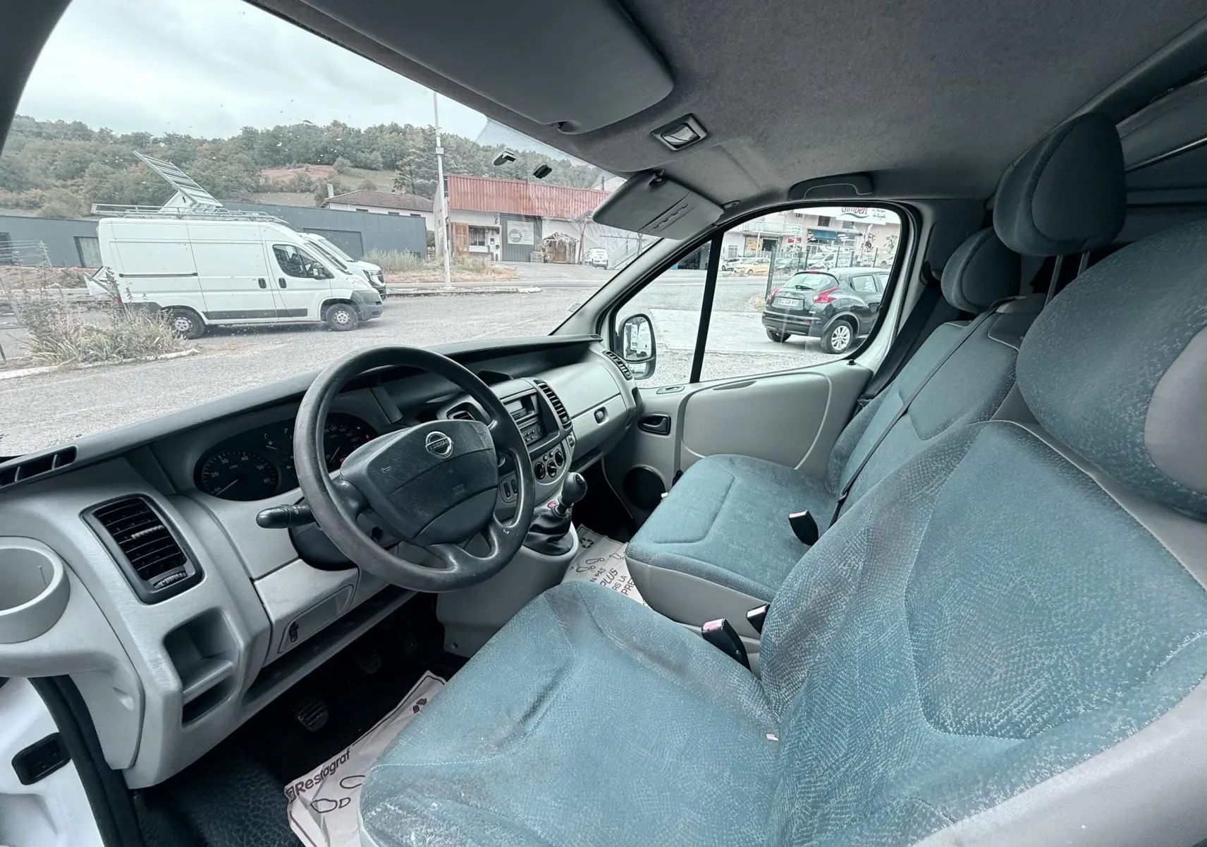 Vue intérieure côté conducteur du Nissan Primastar 1.9 DCI 100 de 2005 avec sièges tissu gris-bleu et volant cuir noir.