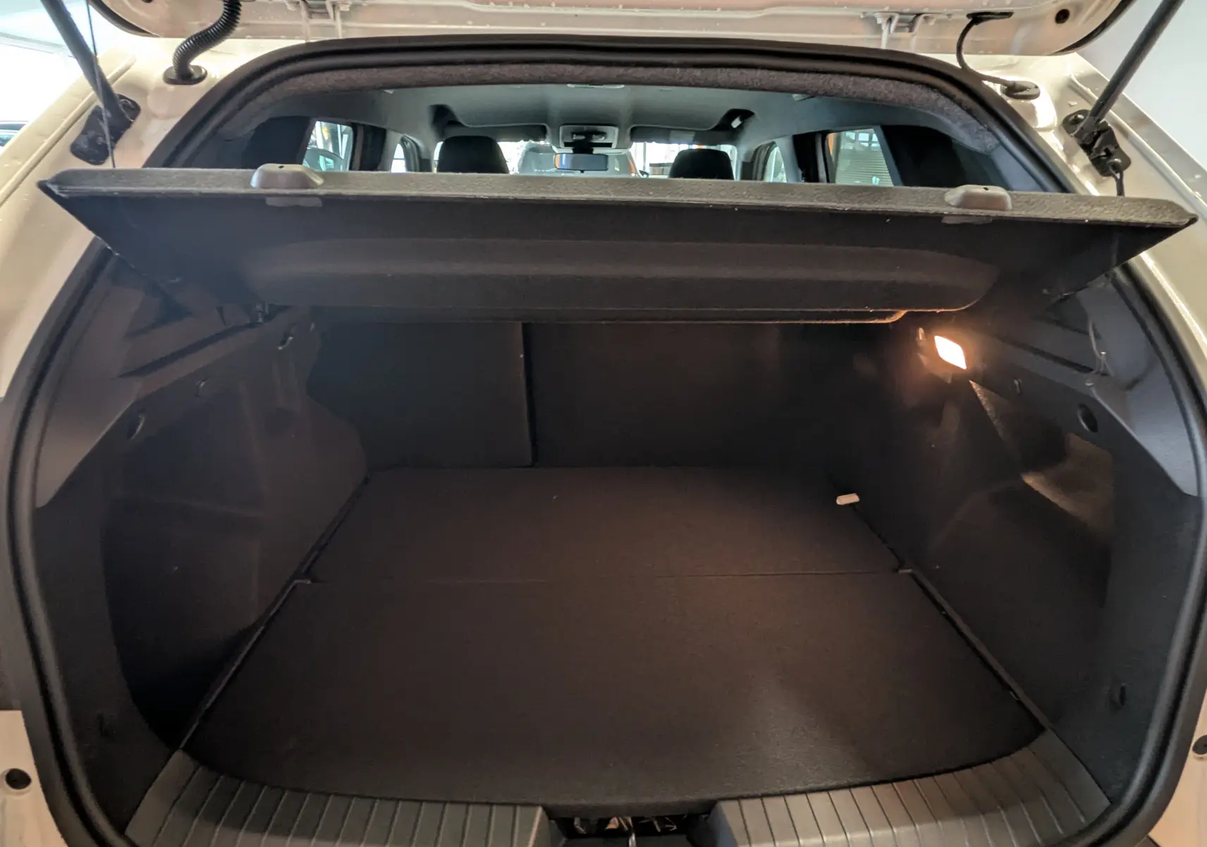 Vue intérieure du coffre ouvert d'un Dacia Duster Hybrid 140 blanc glacier, avec cache-bagages relevé et lumière allumée à droite.
