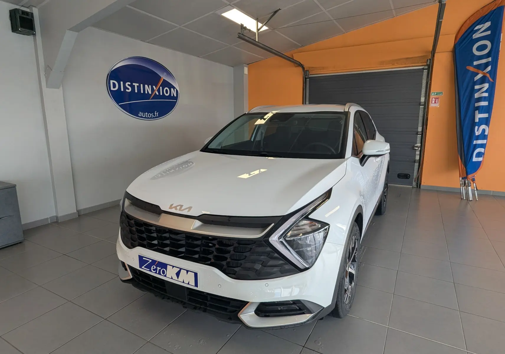 Vue 3/4 avant d'un KIA Sportage blanc 2025 avec calandre noire, jantes alliage bicolores et barres de toit visibles en intérieur showroom.