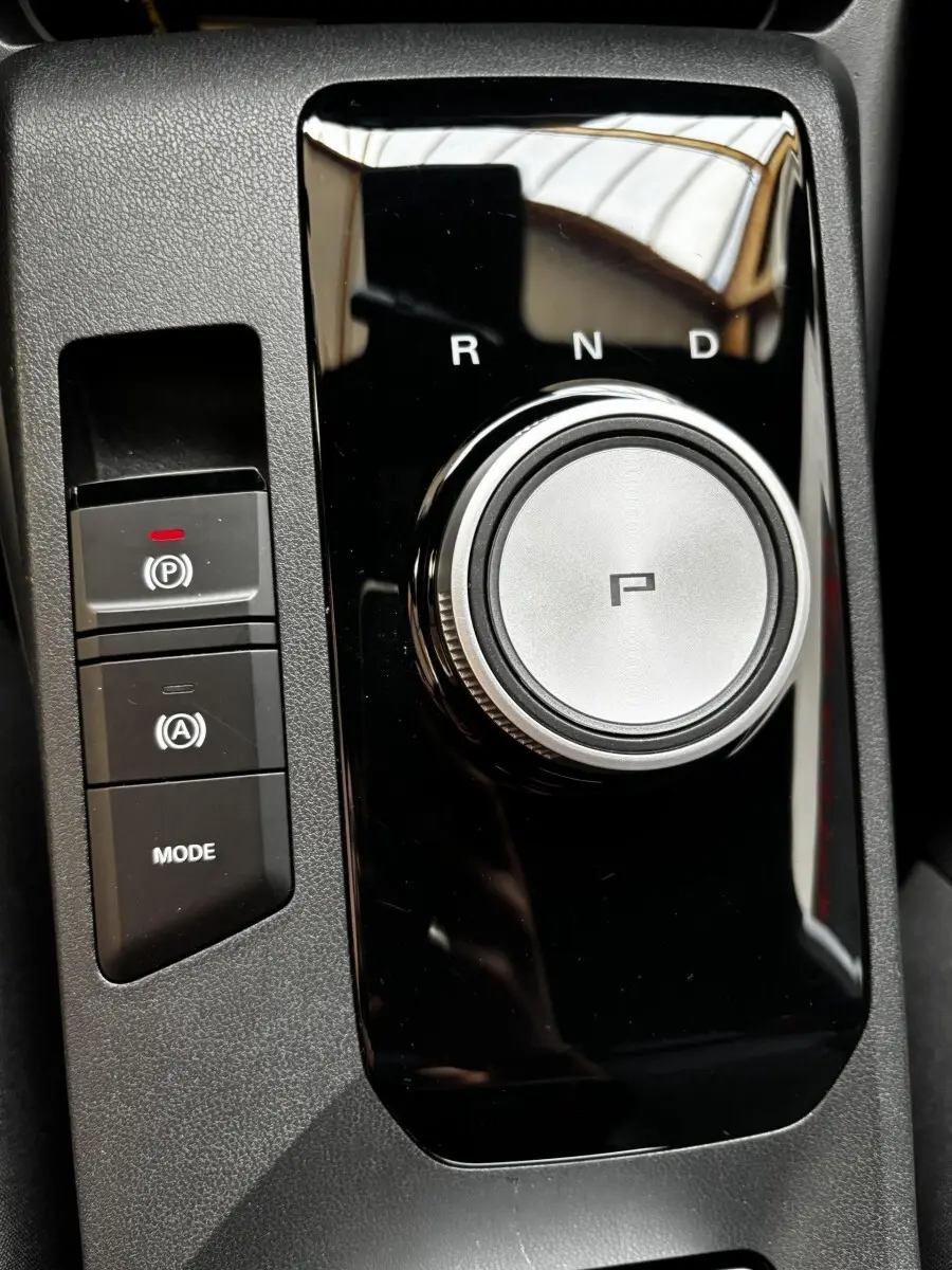 Vue rapprochée du sélecteur rotatif de boîte automatique et des boutons de frein à main et mode sur console noire brillante d’une MG3 gris clair.