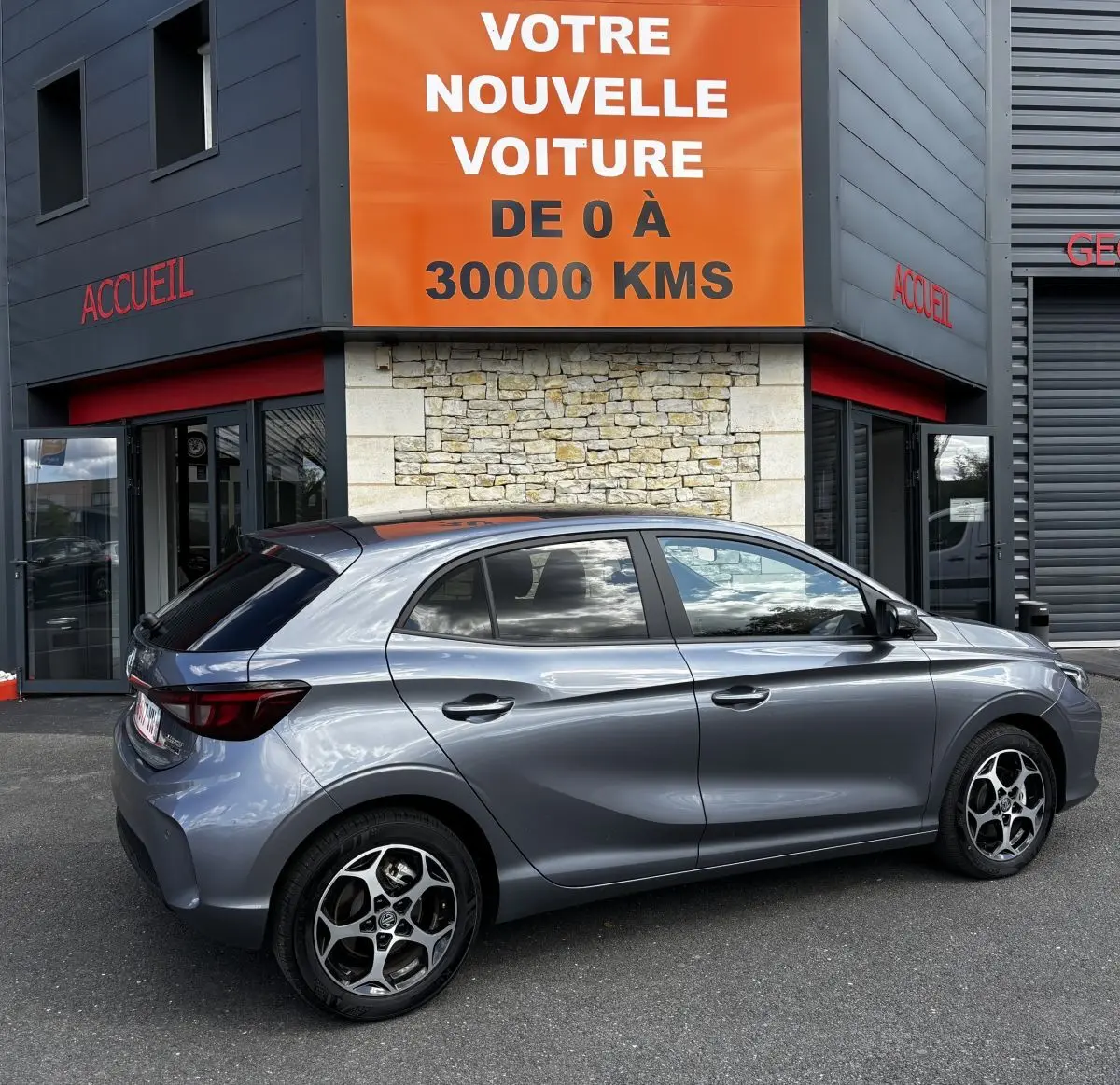 Vue latérale droite d'une MG3 gris clair 2025 garée devant un bâtiment avec panneau orange "Votre nouvelle voiture".