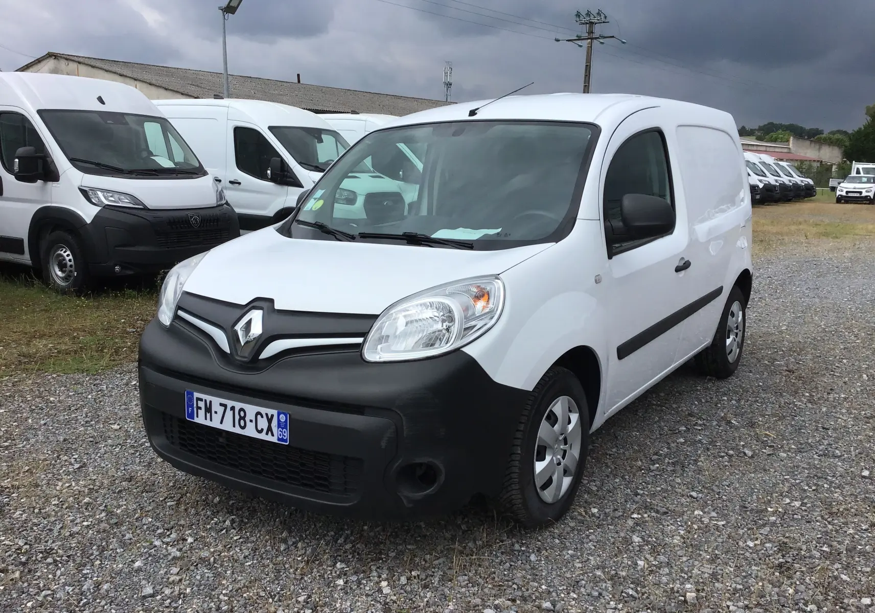 Renault Kangoo blanc en 3/4 avant droit avec pare-chocs noir mat sur un terrain gravillonné sous ciel nuageux.