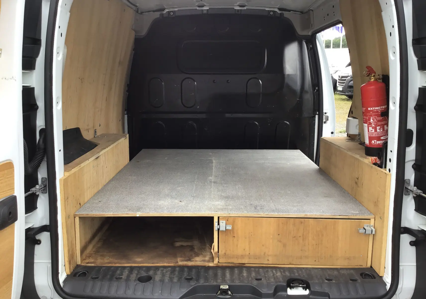 Vue arrière du Renault Kangoo blanc 2019 montrant l'espace de chargement aménagé avec bois et extincteur fixé à droite.