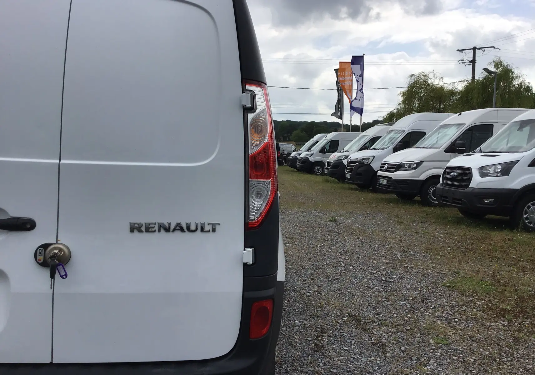 Vue arrière gauche d'un Renault Kangoo blanc 2019 avec serrure et clé visibles, stationné parmi d'autres utilitaires blancs.