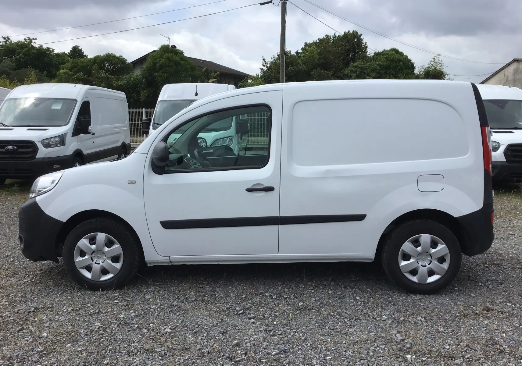 Profil côté gauche d'un utilitaire Renault Kangoo blanc 2019 avec porte latérale et protections noires sur un sol gravillonné.
