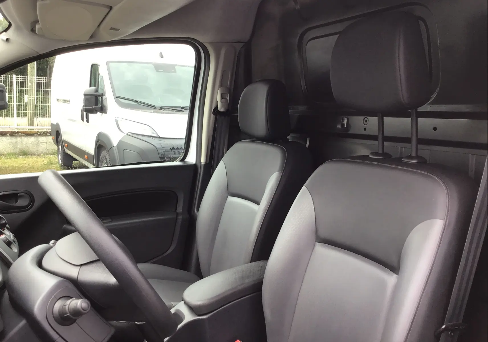 Vue intérieure côté conducteur du Renault Kangoo blanc, montrant les sièges avant en cuir gris et la cloison arrière noire.