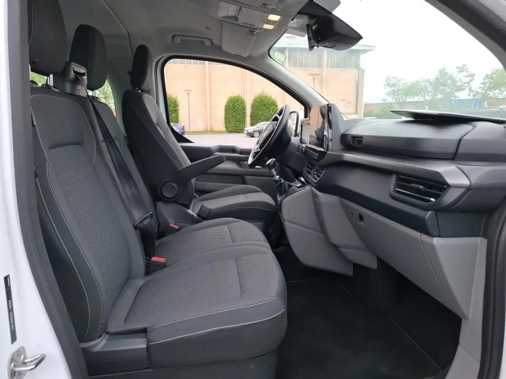 Vue intérieure côté conducteur du Ford Tourneo Custom blanc 2024, montrant sièges tissu gris et tableau de bord moderne.