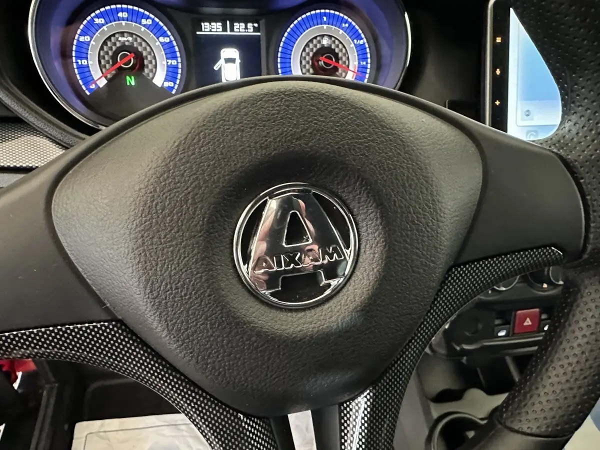 Vue rapprochée du volant noir avec logo Aixam, tableau de bord bleu et écran tactile partiel à droite.