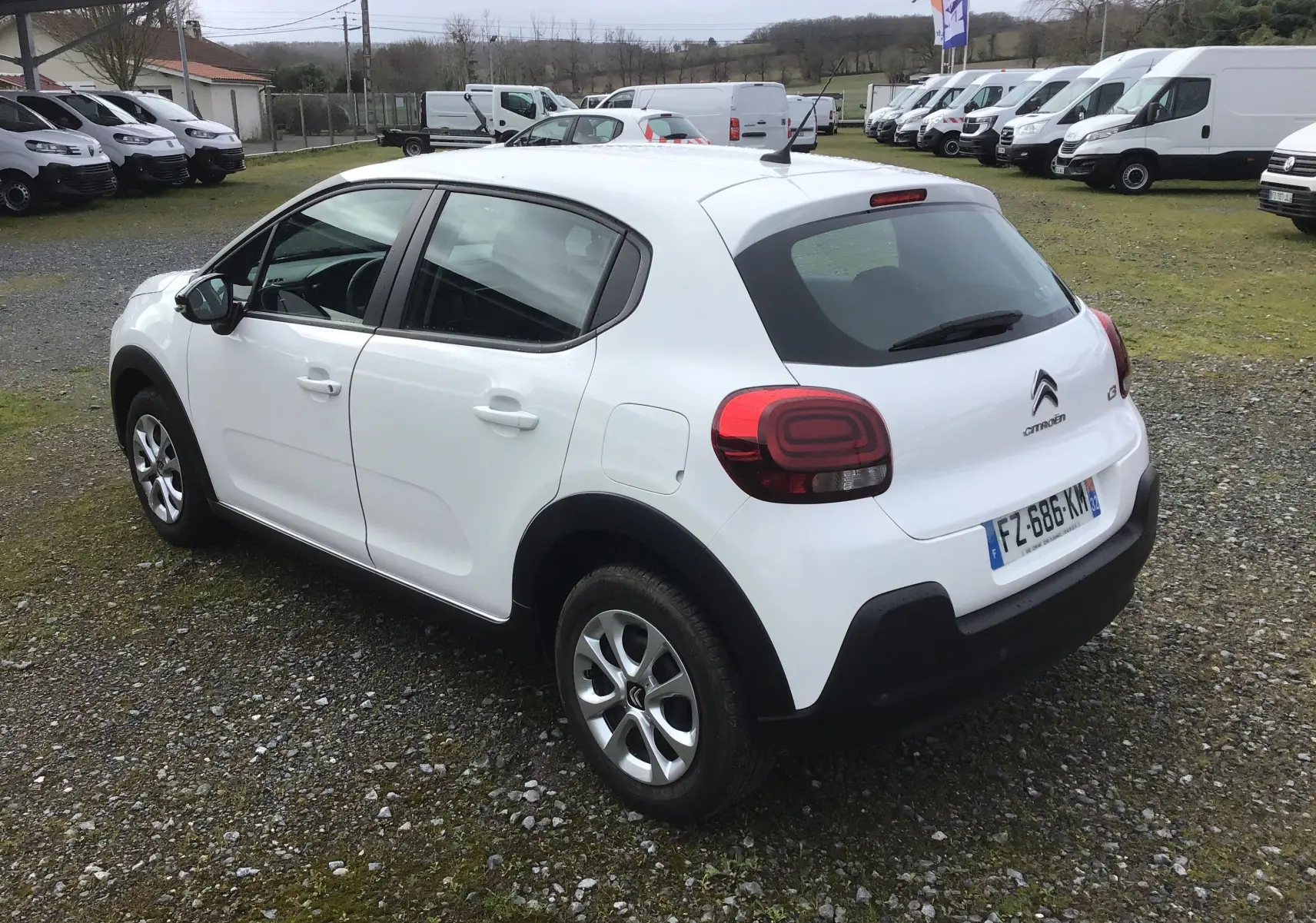Citroën C3 blanche vue 3/4 arrière droit, avec feux arrière arrondis et protections noires sur les bas de caisse.