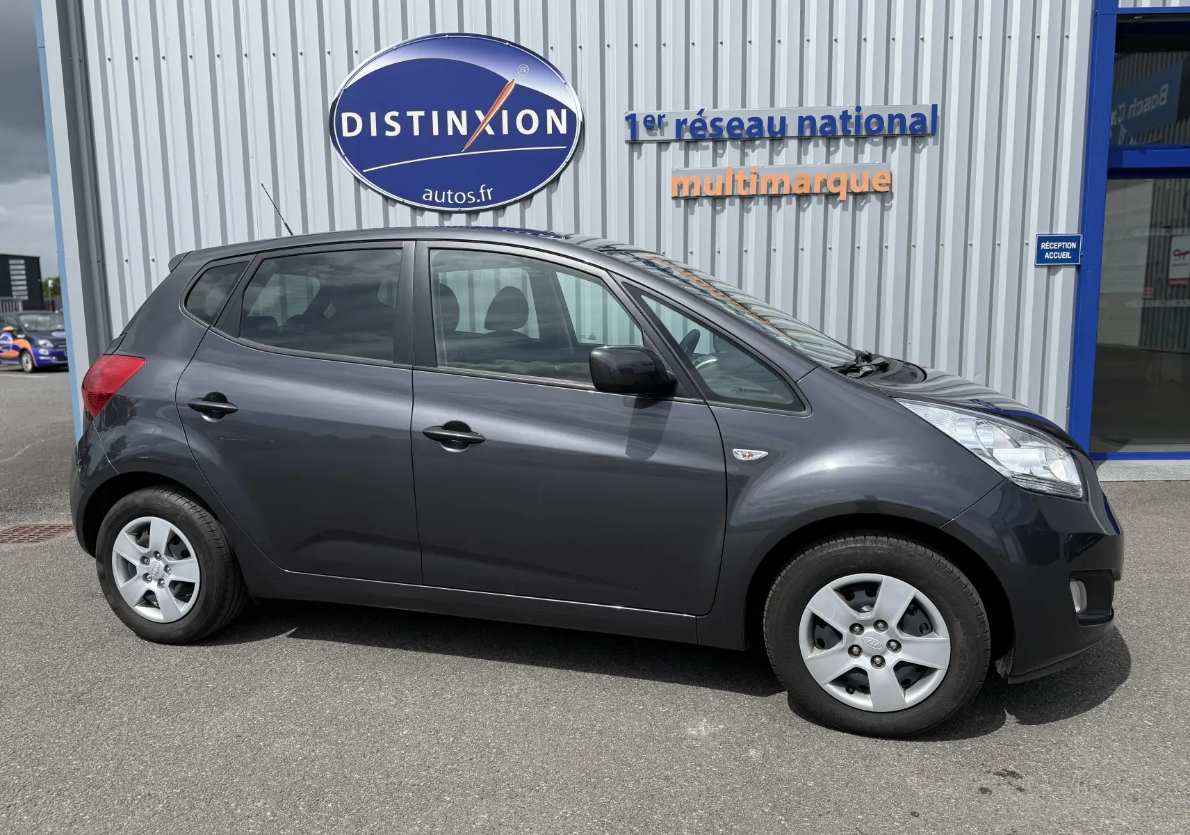 Vue de profil côté gauche d'une KIA VENGA 1.4 i 90 ACTIVE gris foncé garée devant un bâtiment commercial.