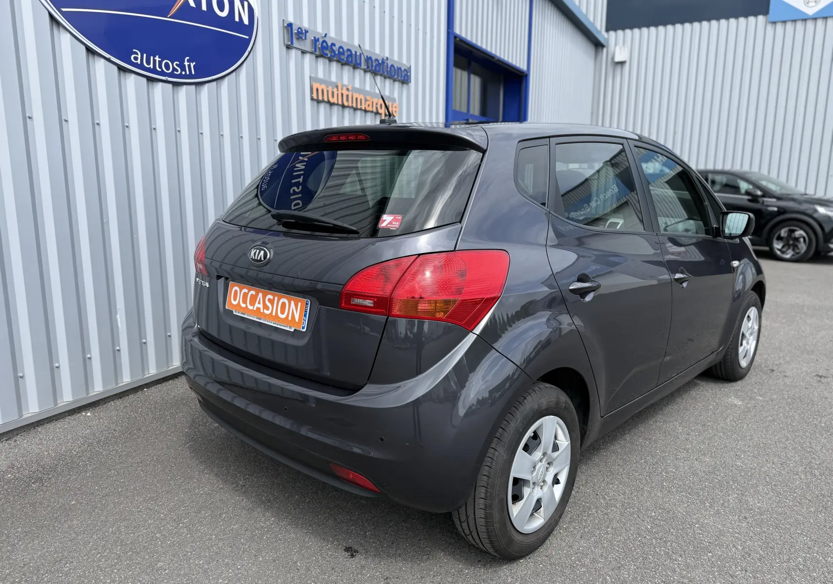 Vue 3/4 arrière droite d'une KIA VENGA gris foncé de 2013 avec plaque "OCCASION" sur un parking extérieur.