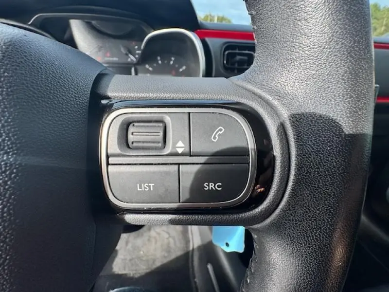 Gros plan sur les commandes audio au volant cuir d'une Citroën C3 2017, avec tableau de bord rouge Aden en arrière-plan.