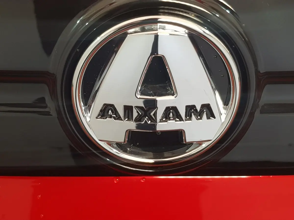 Gros plan sur le logo chromé AIXAM sur fond noir et rouge vif de la carrosserie d'une Aixam Ambition 2026.