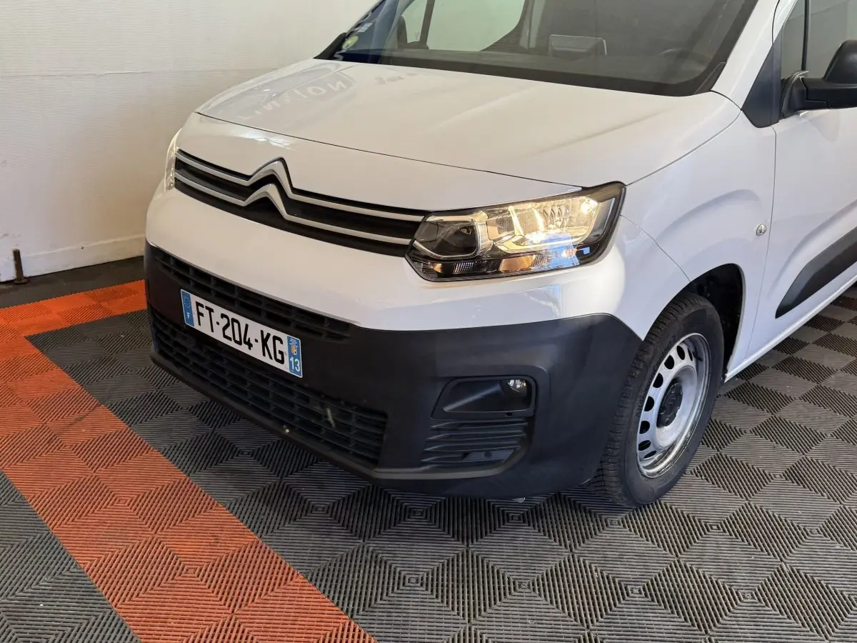 Vue 3/4 avant droit du Citroën Berlingo blanc avec pare-chocs noir et phares allumés sur sol carrelé.