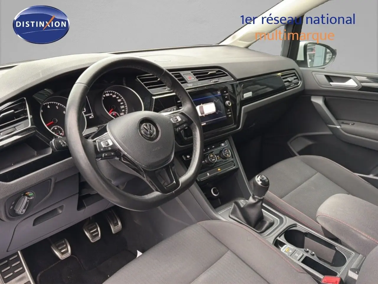 Vue intérieure côté conducteur du Volkswagen Touran 2018, montrant le volant, la console centrale et les sièges tissu gris.