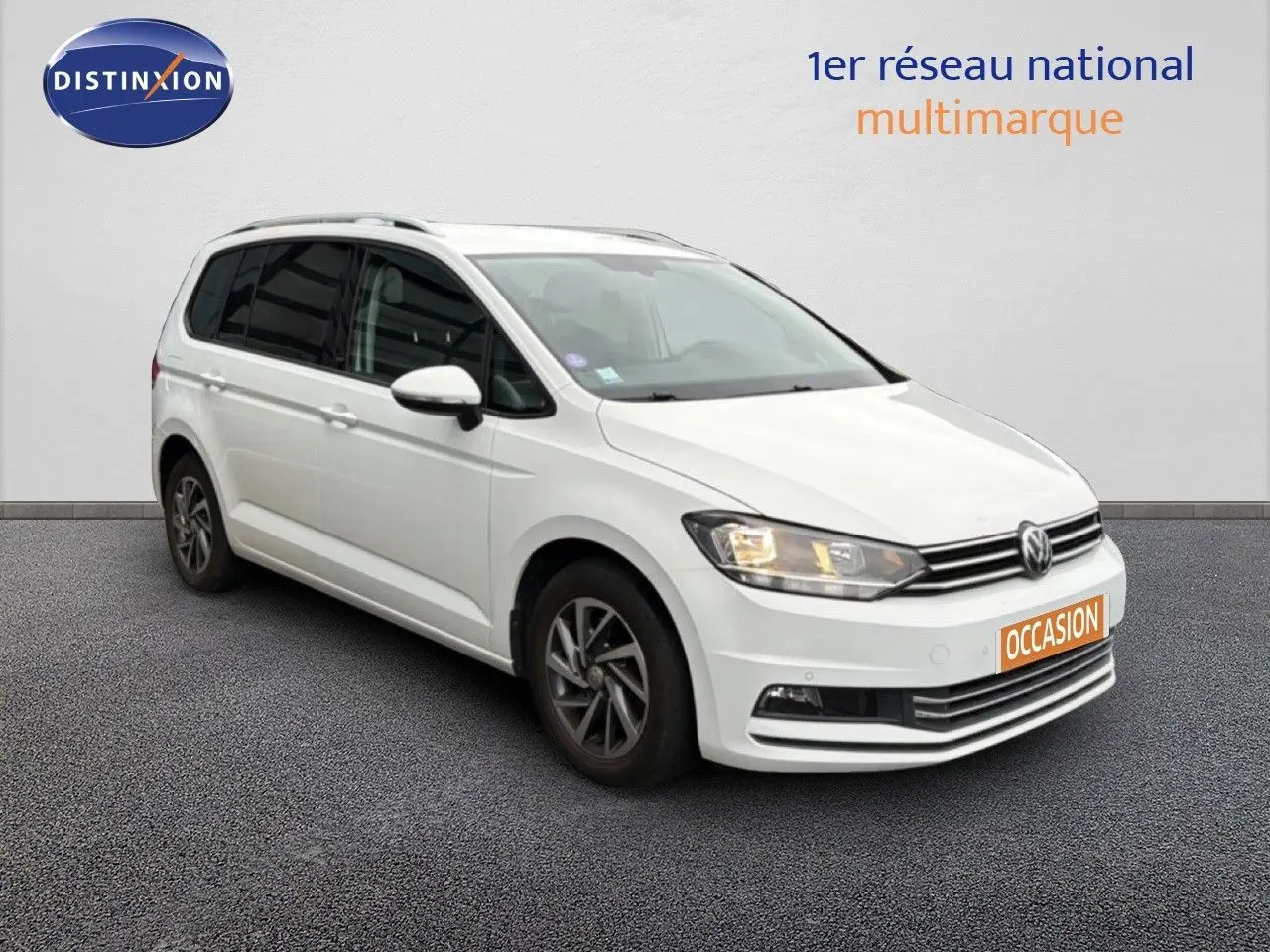 Volkswagen Touran blanc vu en 3/4 avant droit avec phares allumés et jantes bi-ton.