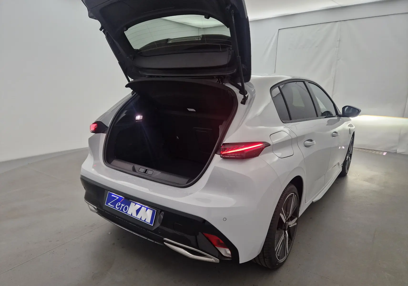 Vue 3/4 arrière droite d'une Peugeot 308 blanche Okenite 2025 avec coffre ouvert et feux arrière allumés.