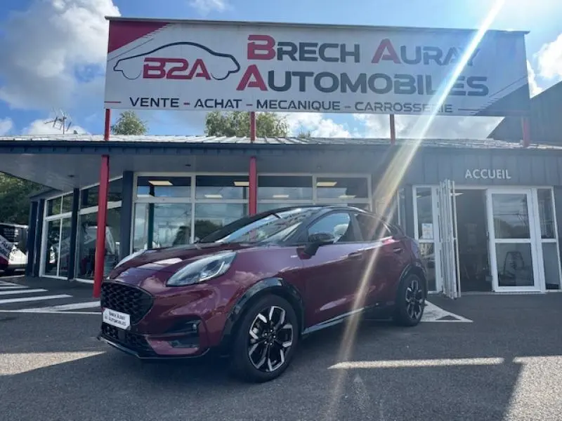 Ford Puma rouge Vivid Ruby en 3/4 avant droit devant un garage avec enseigne Brech Aura Automobiles