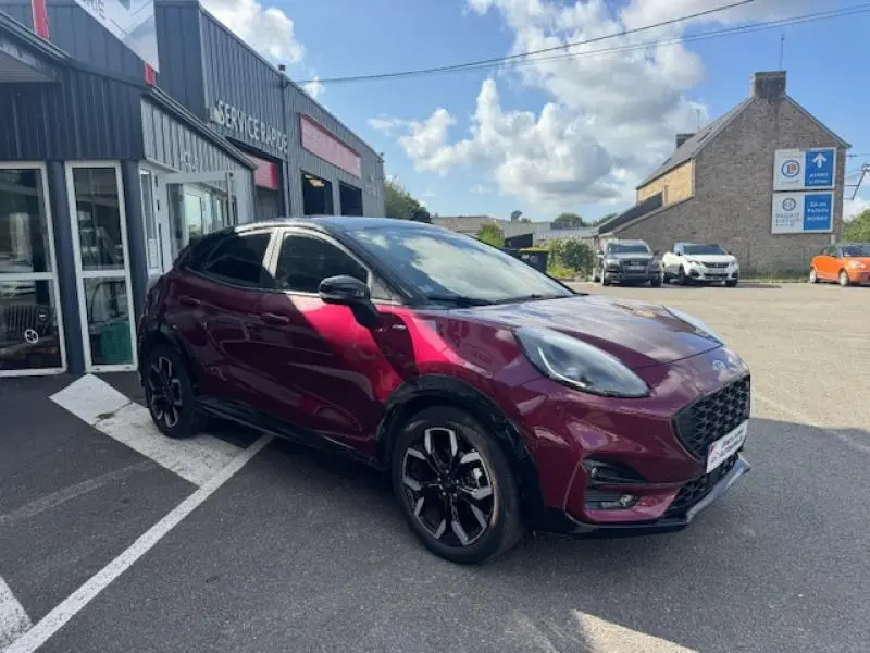 Ford Puma rouge vif vue 3/4 avant droit, avec jantes noires et toit noir brillant, stationnée devant un garage.