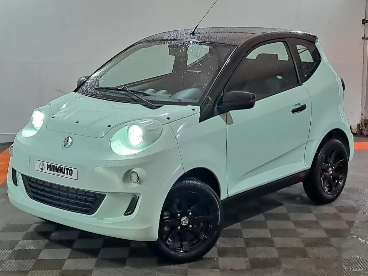 Vue 3/4 avant d'une Aixam Minauto E chic 2026 vert clair avec toit noir et jantes noires, phare avant allumé.