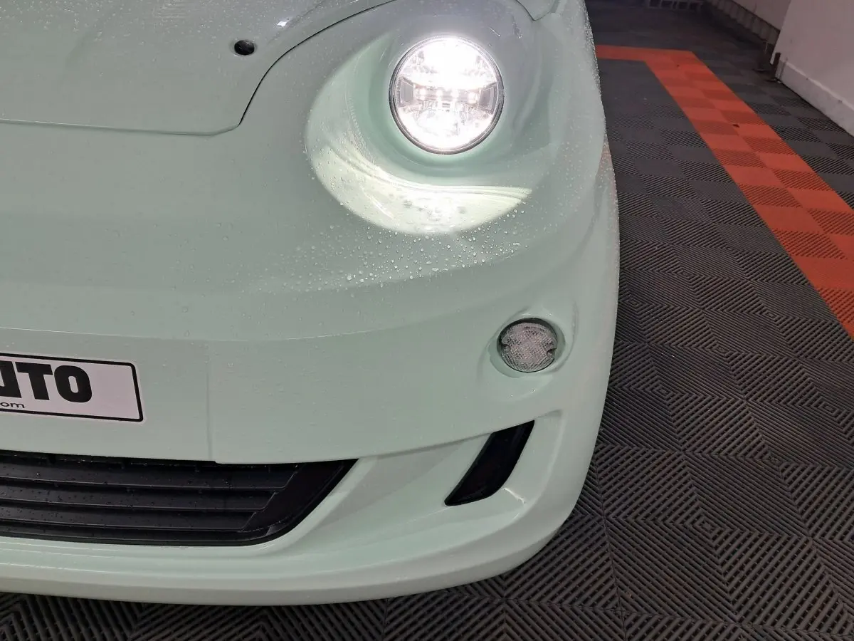 Gros plan sur l'avant droit vert clair de l'AIXAM MINAUTO E chic 2026 avec phare allumé et gouttes d'eau visibles.