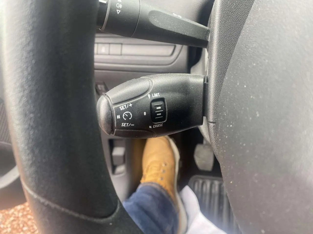 Gros plan sur la commande de régulateur de vitesse au volant d'une Peugeot 208 gris shark, avec pédale et chaussure visible.