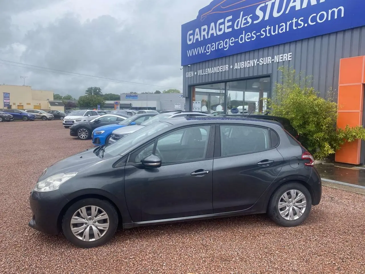 Profil côté gauche d'une Peugeot 208 gris shark 2013 garée devant un garage avec jantes acier et poignées noires.