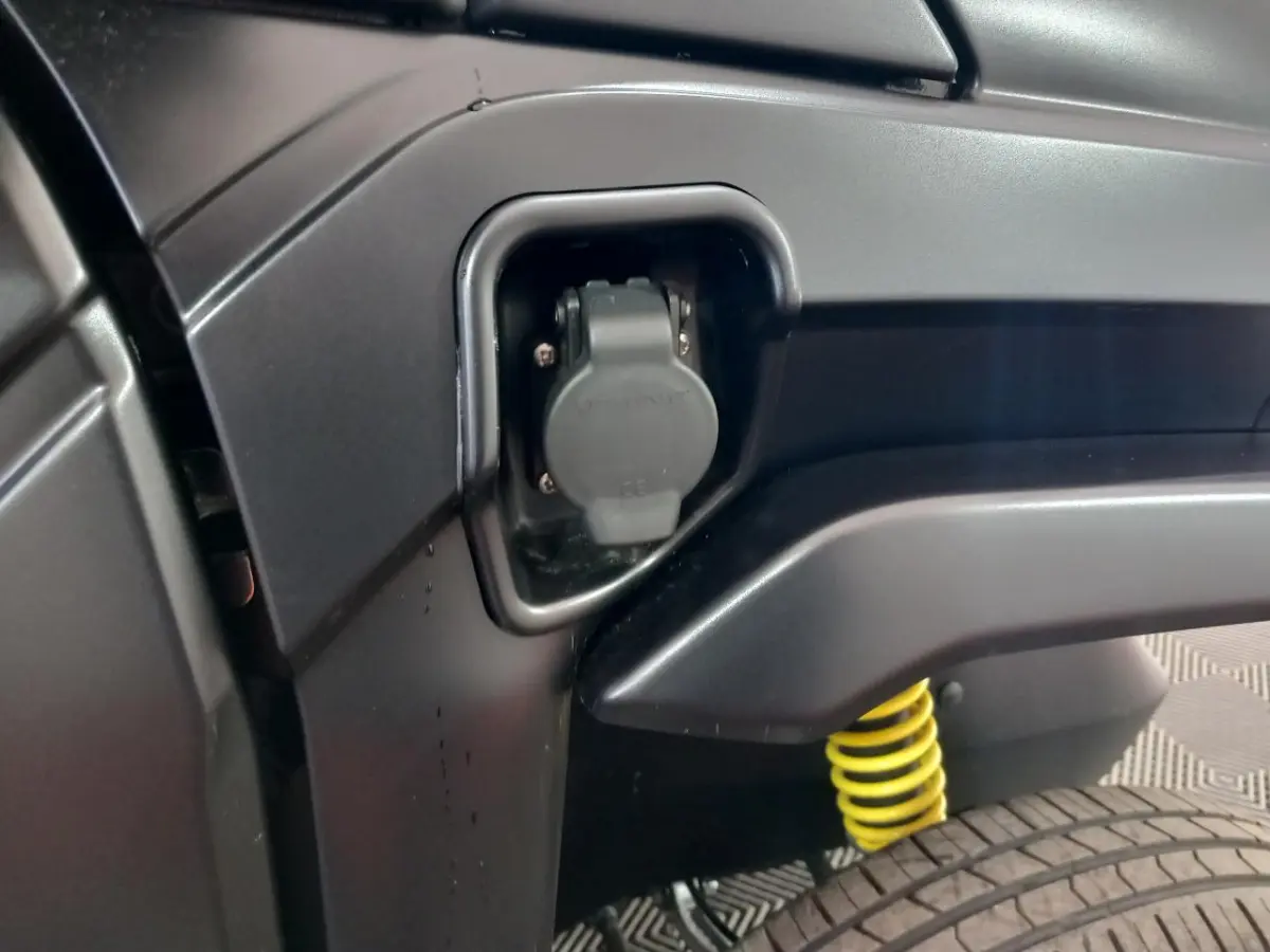 Détail de la prise de recharge électrique côté gauche d'une Aixam Mega E-Scouty Evo bleu, avec ressort de suspension jaune visible.