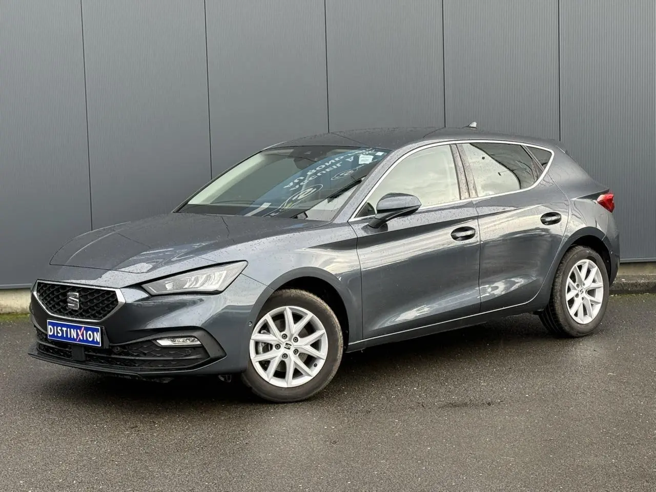 SEAT Leon 1.5 TSI 115 Style gris magnétique en 3/4 avant droit, jantes alliage 16 pouces visibles, stationnée sur sol humide.