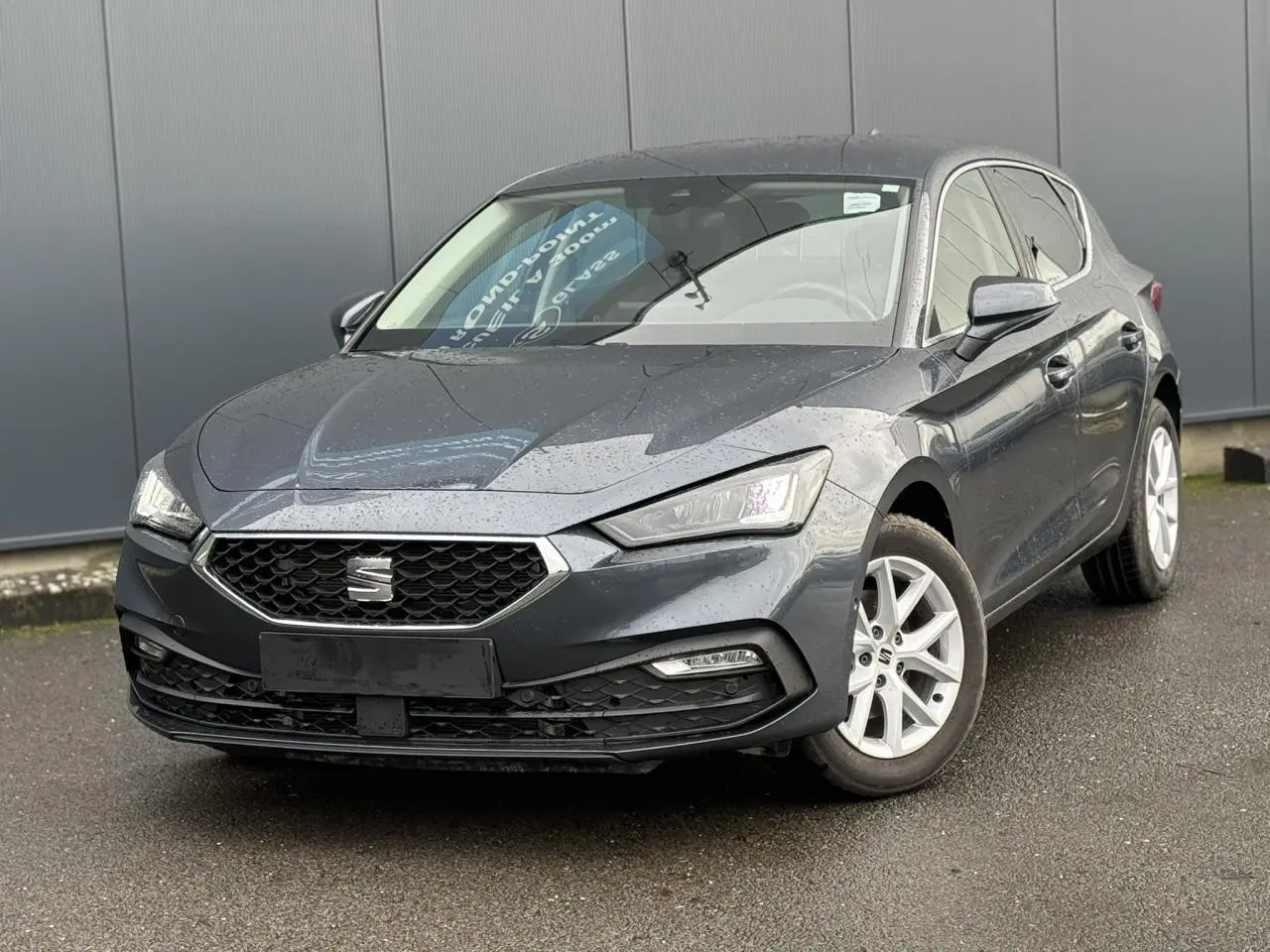 SEAT Leon 1.5 TSI gris magnétique vue 3/4 avant droit avec jantes alliage blanches sur sol humide.