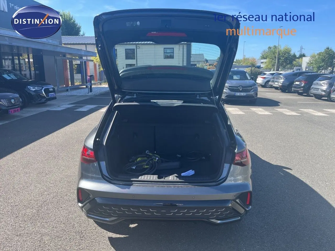 Vue arrière d'une Audi A3 Sportback gris Daytona avec coffre ouvert montrant câbles de recharge dans un parking extérieur.