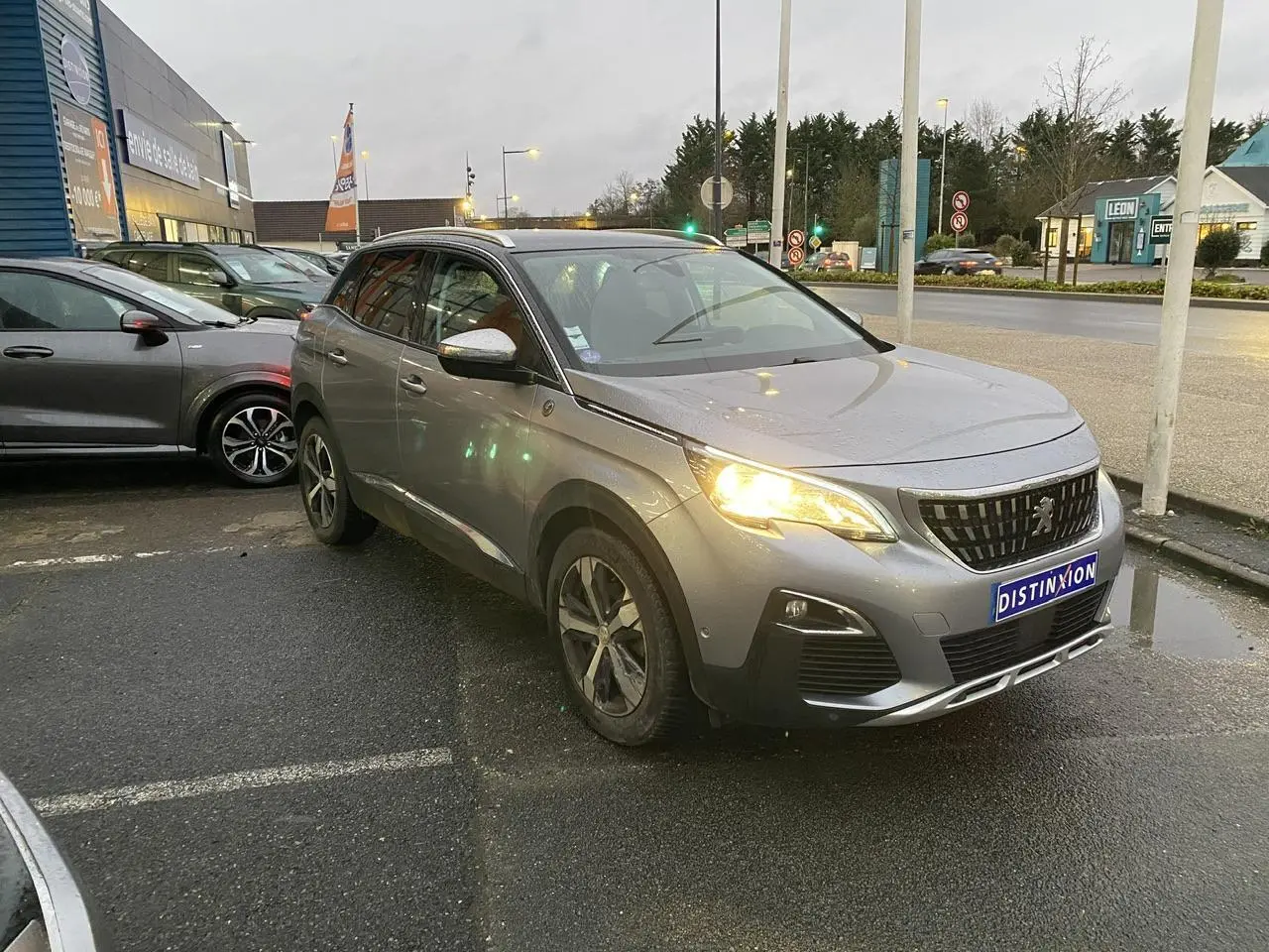 Peugeot 3008 gris Artense vue 3/4 avant droit avec phares allumés sur parking urbain.
