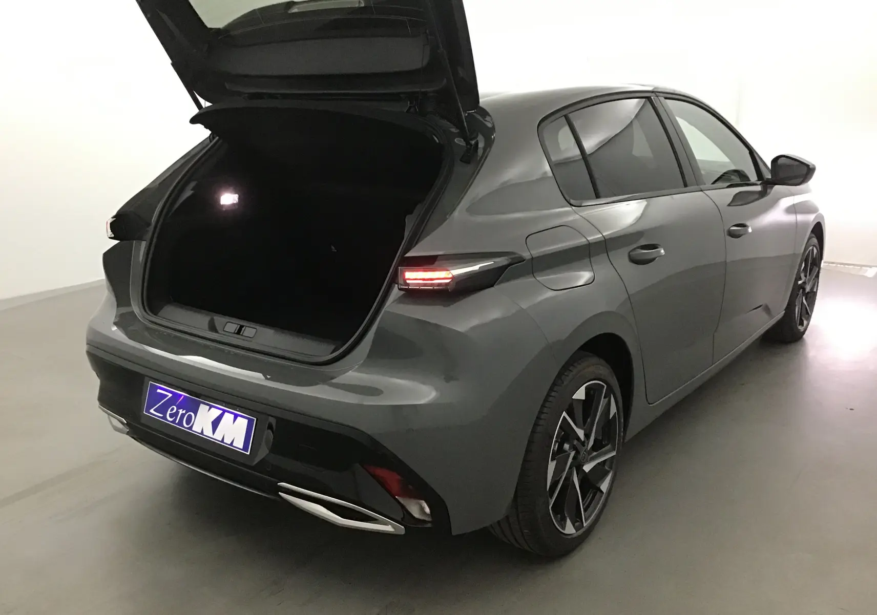 Vue 3/4 arrière droite d'une Peugeot 308 gris Sellenium avec coffre ouvert et feux arrière allumés.