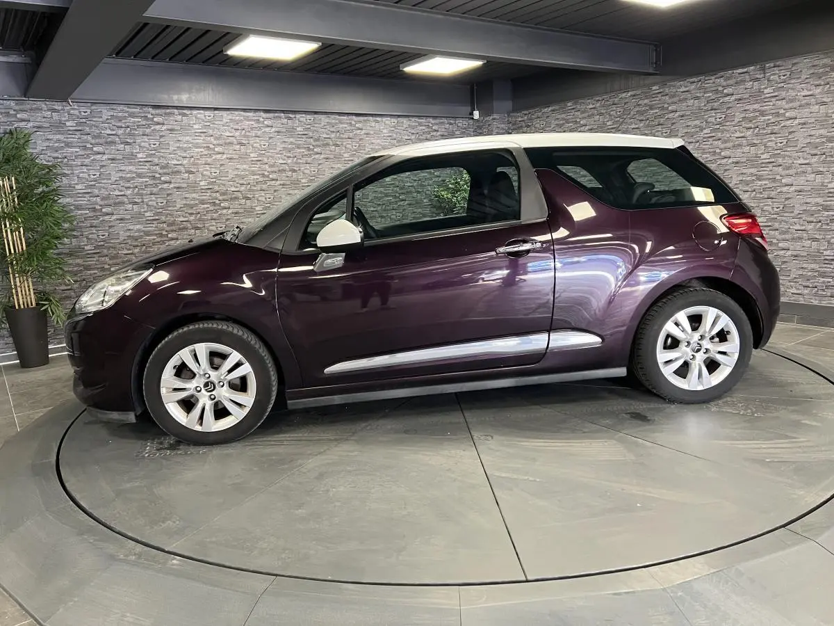 Profil côté gauche d'une DS3 violet avec toit blanc, jantes alliage et poignées chromées en intérieur showroom.