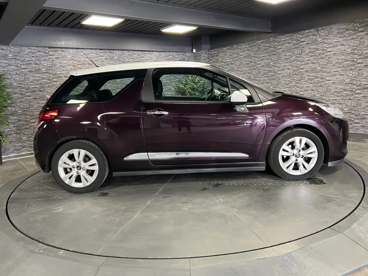 Profil côté gauche d'une DS3 violet avec toit blanc et jantes alliage dans un showroom intérieur.
