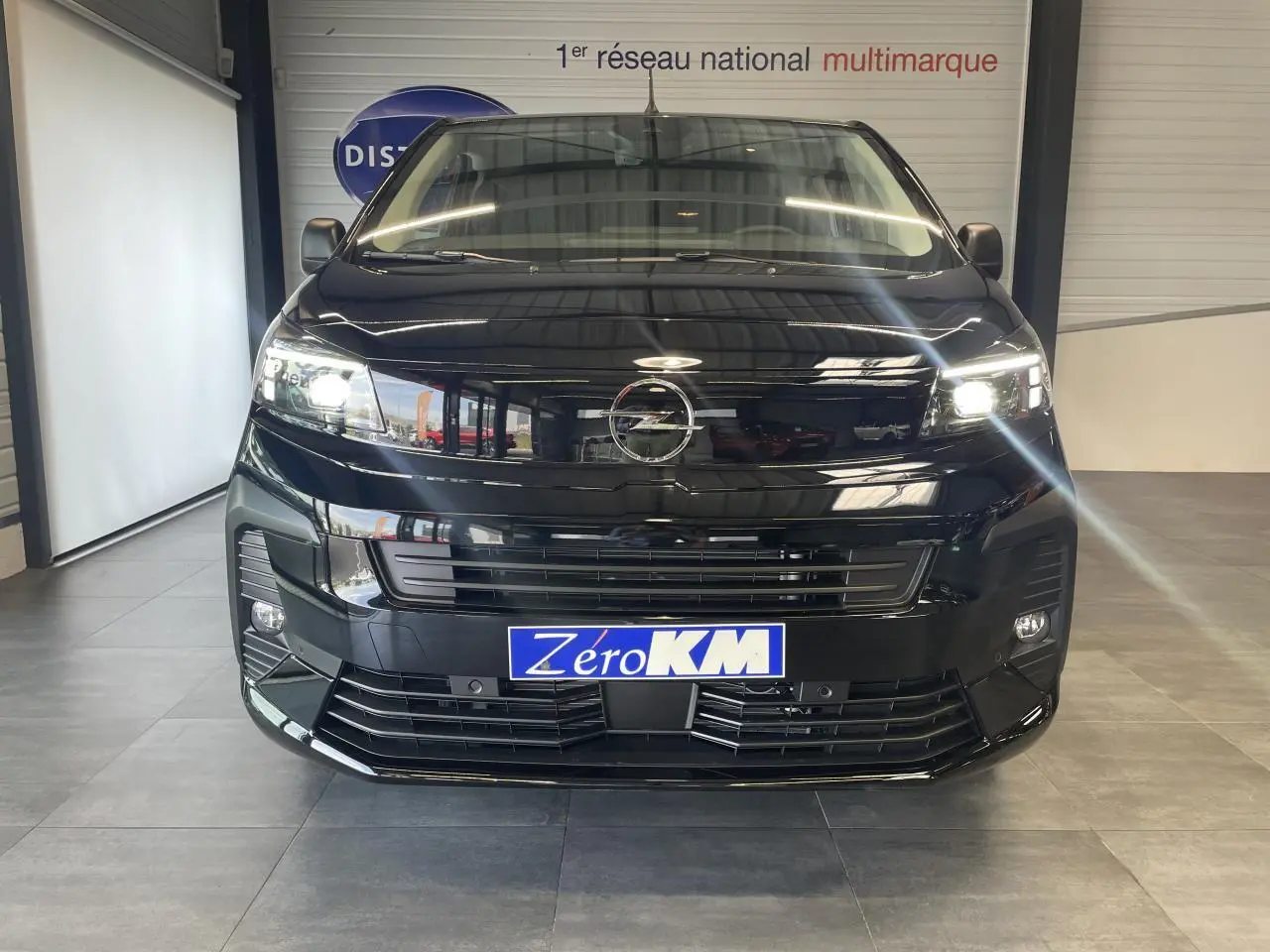 Vue frontale d'un Opel Vivaro noir carbone 2025 avec phares allumés et plaque Zéro KM visible.