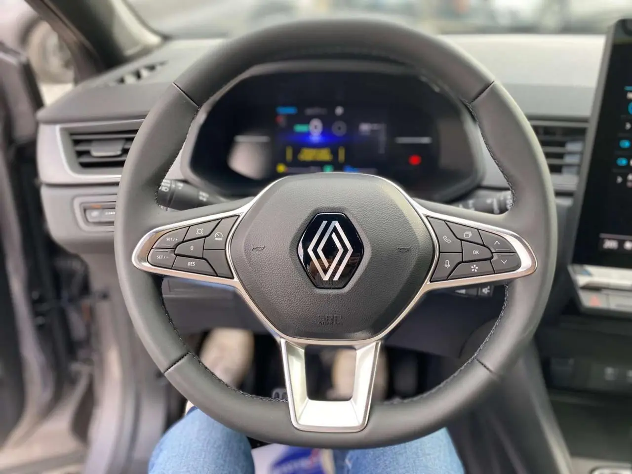 Vue rapprochée du volant multifonction noir avec logo Renault, tableau de bord numérique flou en arrière-plan.