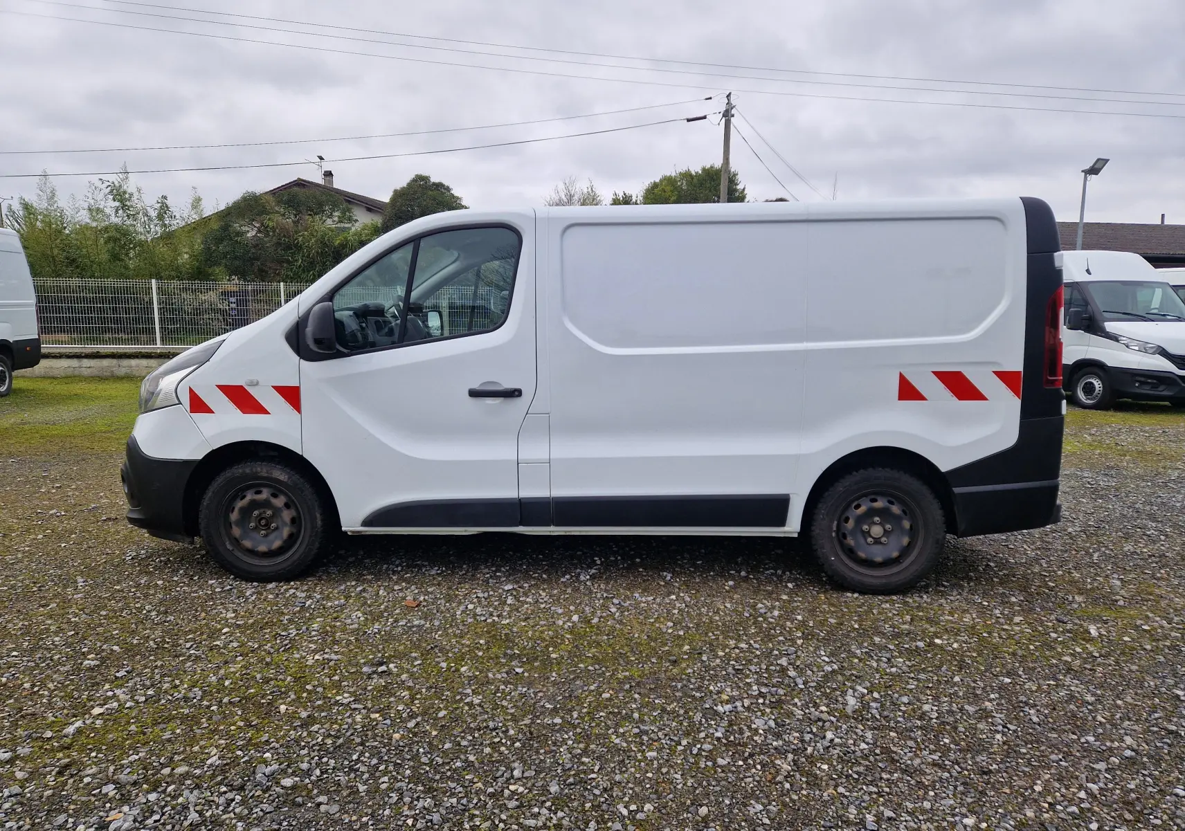 Vue de profil côté gauche d’un Renault Trafic Fourgon blanc avec bandes réfléchissantes rouges sur l’aile avant et l’arrière.