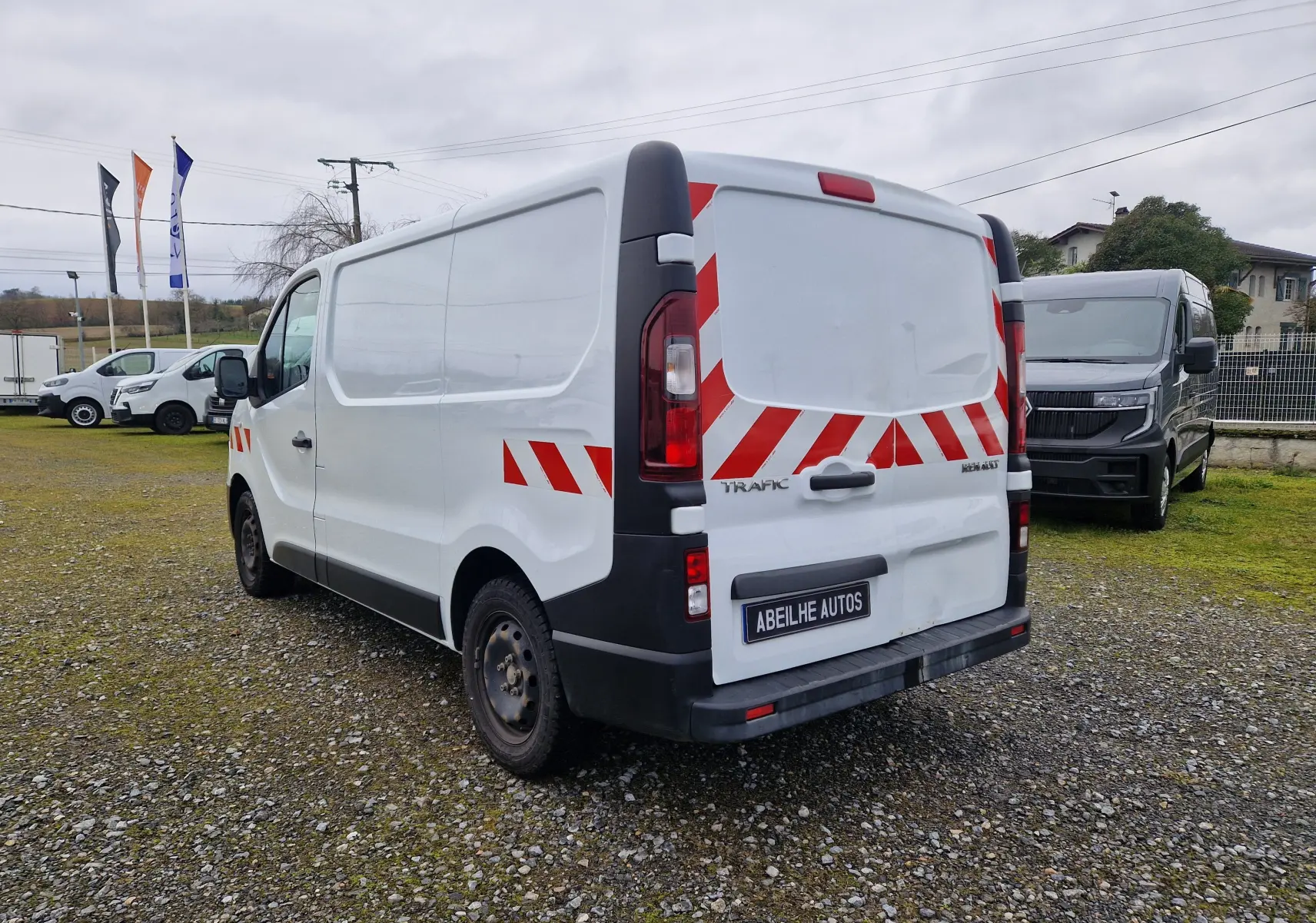 Vue 3/4 arrière droite d’un Renault Trafic blanc avec bandes rouges réfléchissantes sur un terrain gravillonné.