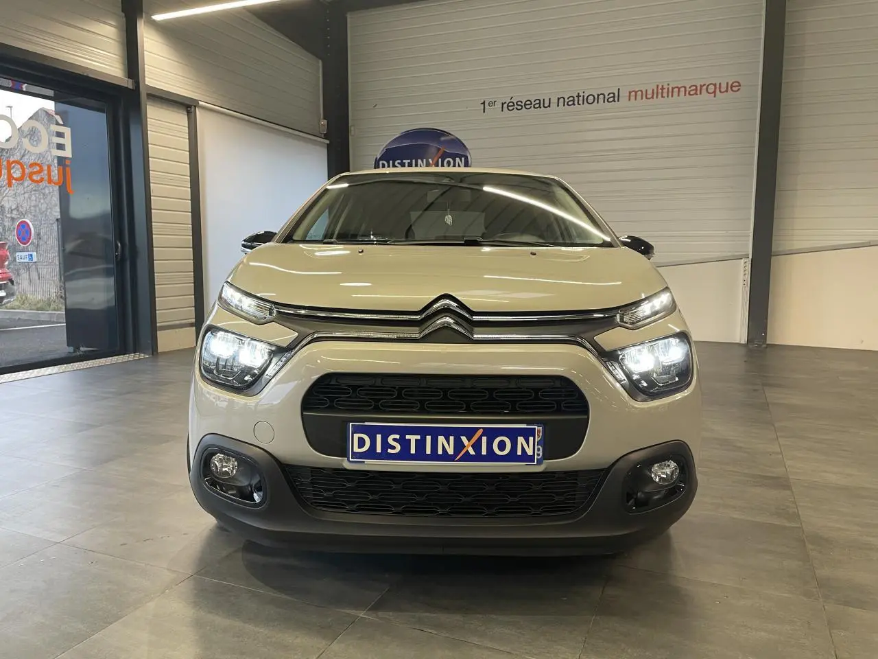 Vue frontale d'une Citroën C3 beige clair 2024 avec feux allumés et calandre noire en showroom.