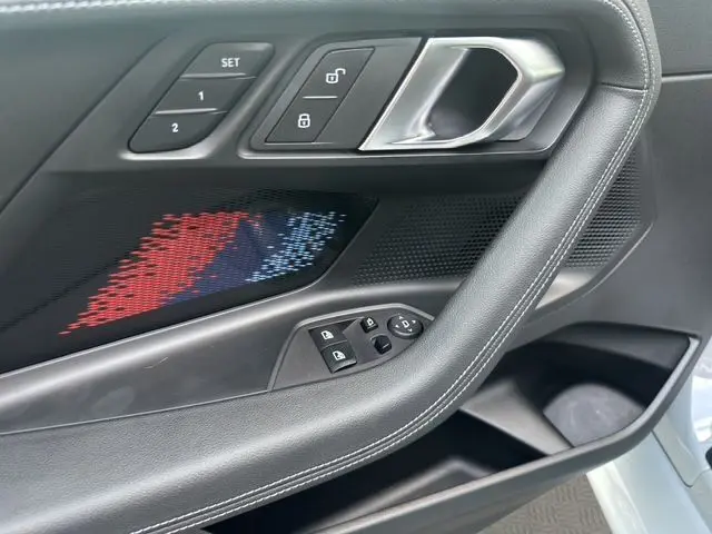 Gros plan sur la porte côté gauche de la BMW M2 G87 gris Brooklyn avec éclairage décoratif tricolore et commandes électriques.