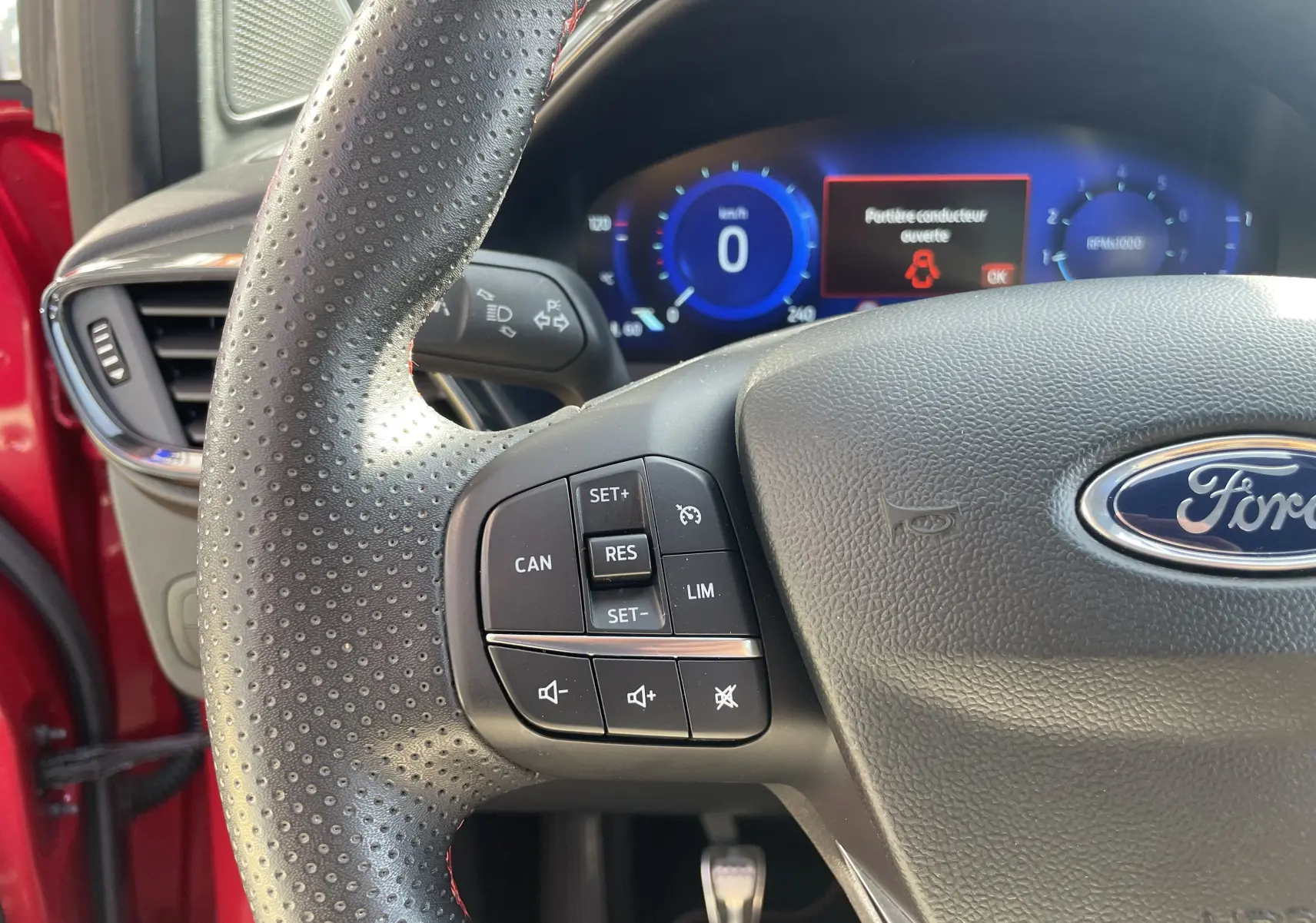 Gros plan sur le volant perforé d'une Ford Puma rouge 2021, avec commandes intégrées et tableau de bord numérique bleu en arrière-plan.
