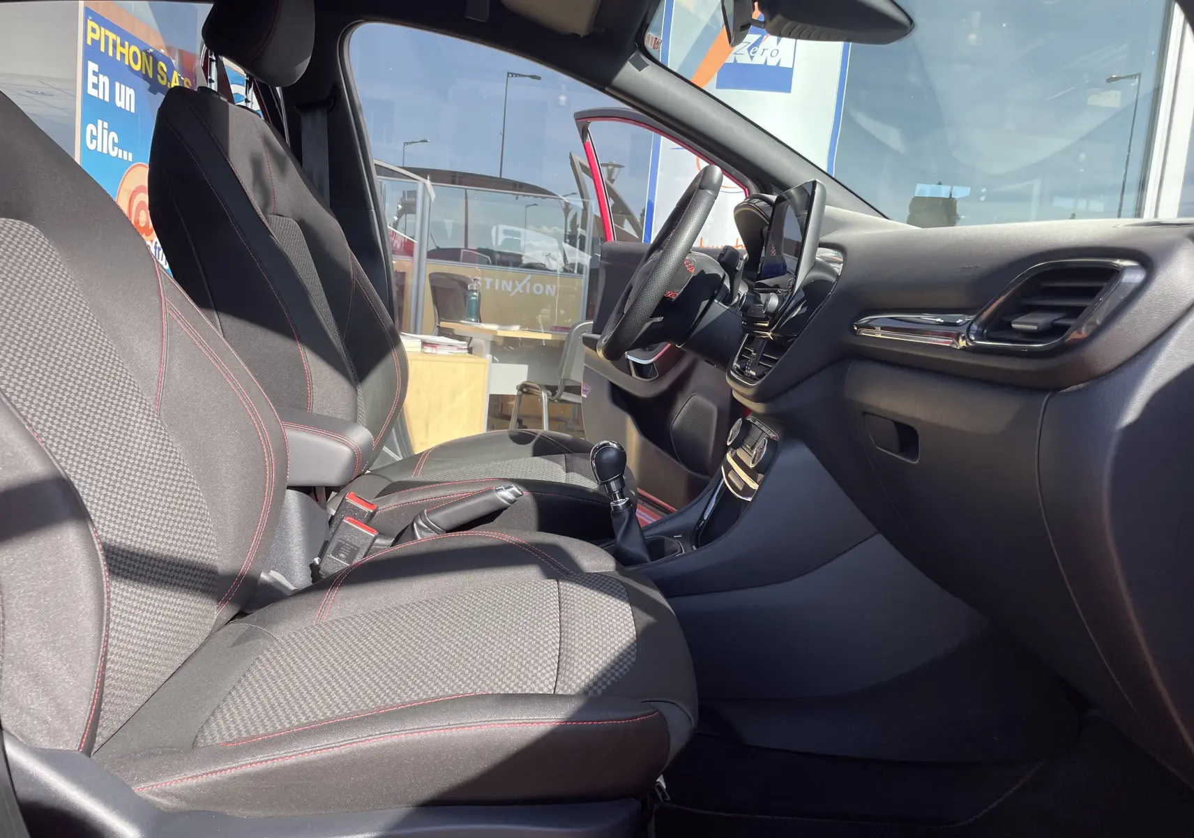 Intérieur du Ford Puma rouge 2021 vu côté conducteur, sièges tissu noir avec surpiqûres rouges et tableau de bord moderne.