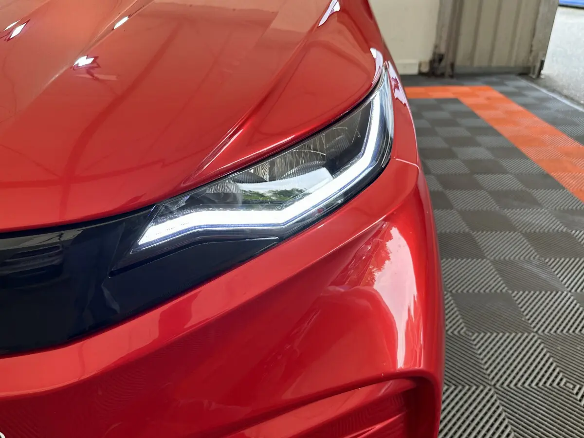 Gros plan sur le phare avant droit allumé d'une Aixam Ambition E City Pack rouge, soulignant ses lignes modernes.