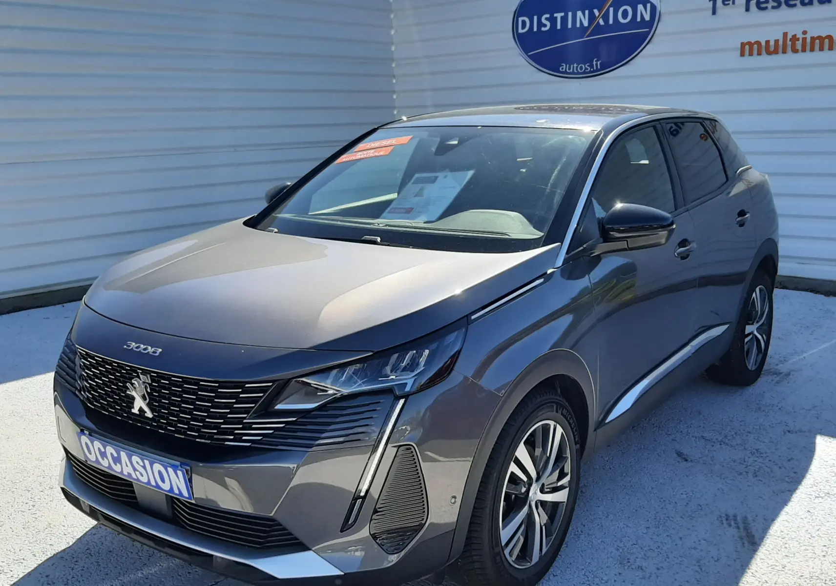 Peugeot 3008 gris platinium en 3/4 avant droit, avec calandre noire et feux LED distinctifs, stationné devant un mur blanc.