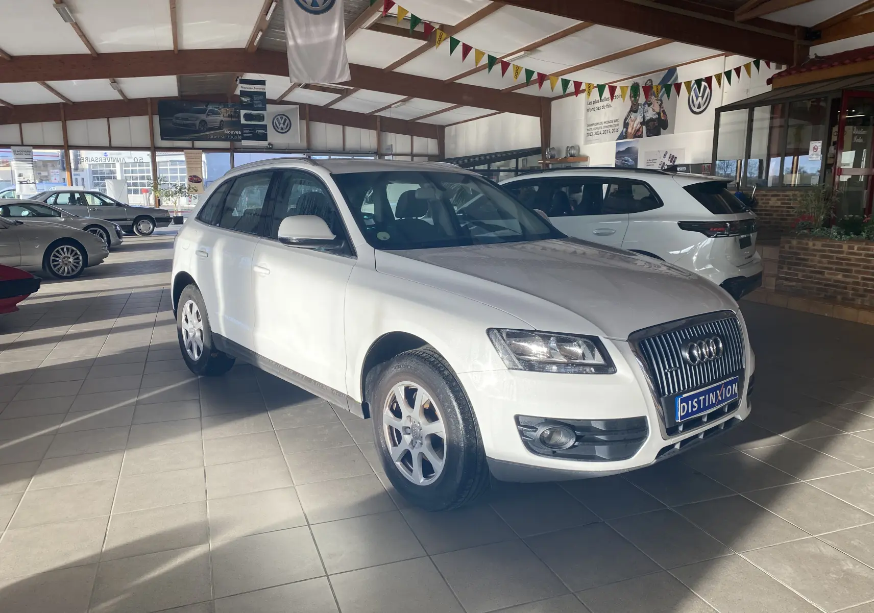 Audi Q5 blanc vue 3/4 avant droit dans un showroom avec plaques "Distinxion" et drapeaux colorés suspendus.