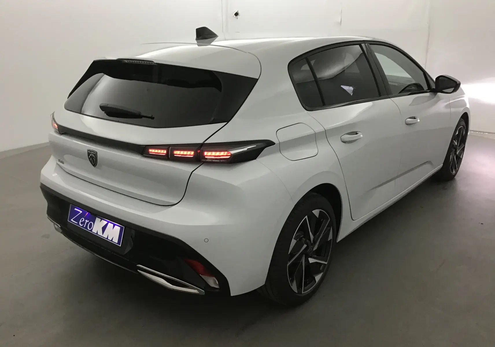 Vue 3/4 arrière droite d'une Peugeot 308 blanche Okenite avec feux arrière LED allumés et jantes alliage noires.